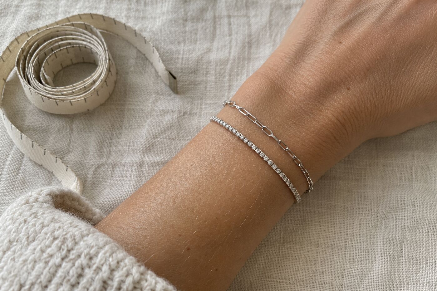 Pols met een dunne zilveren schakelarmband en een smalle tennisarmband, met een opgerold meetlint ernaast